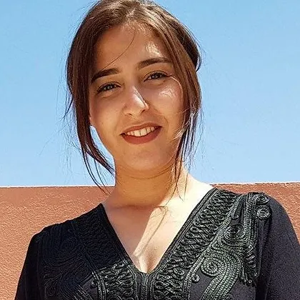 A person smiles in a black embroidered top, standing against a vibrant terracotta wall with the blue Tunisian sky above, capturing the essence of an Experience Tunisia tour. A person smiles in a black embroidered top, standing against a vibrant terracotta wall with the blue Tunisian sky above, capturing the essence of an Experience Tunisia tour.