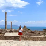 A woman in a red skirt stands by ancient ruins with towering columns, overlooking the vast sea under a cloud-dotted blue sky, capturing the essence of an unforgettable Experience Tunisia.