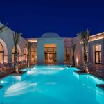 Modern villa exterior with an illuminated pool at night, reminiscent of Tunisia's architectural charm. The design features arched windows and palm trees lining the poolside, creating a serene escape for those looking to experience Tunisia's enchanting ambiance right at home.