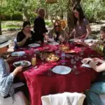 Eight people are seated around a red-clothed table outdoors, sharing a meal surrounded by trees and greenery on a sunny day while in tour in Tunisia with Xperience Tunisia