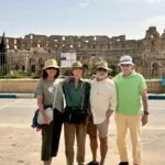 Four adults stand together in front of an ancient Roman amphitheater under a clear sky, posing for a group photo while embracing Tunisia cultural norms during their travel.