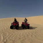 Two people ride red quad bikes on sand dunes under a clear blue sky, adding adventure to their trekking in Tunisia experience.