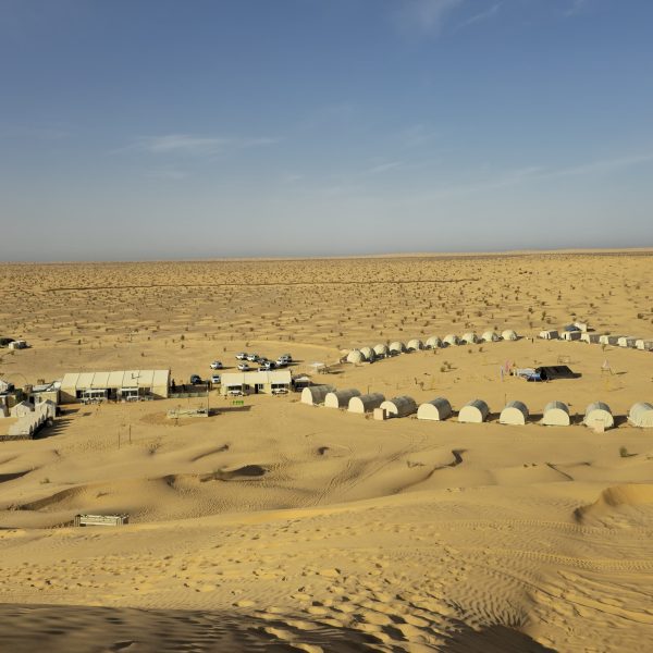 Aerial view of one of the best Sahara Desert Camps in Tunisia, with numerous white tents arranged in an oval shape on sandy terrain under a clear blue sky.